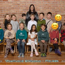Eine Gruppe von Kindern und einer Frau posiert für ein Foto vor einer Ziegelwand mit lächelnden Emojis. Die Kinder sitzen auf Holzstühlen, während die Frau hinter ihnen steht. Das Foto hat einen Wasserzeichen am unteren Rand.