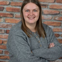 A woman with brown hair wearing a gray sweater is smiling and posing for a photo with her arms crossed in front of a brick wall.