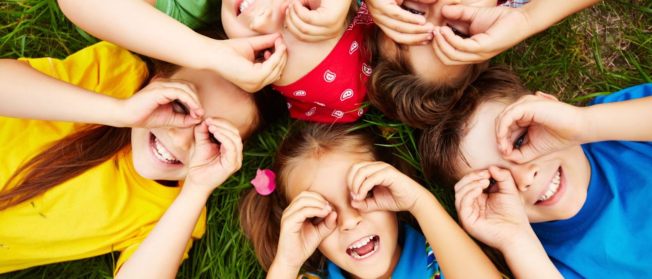 Mehrere Kinder liegen auf einem Grasfeld und lächeln. Sie machen lustige Gesichter und haben ihre Hände um die Augen gelegt, möglicherweise spielen sie ein Spiel.