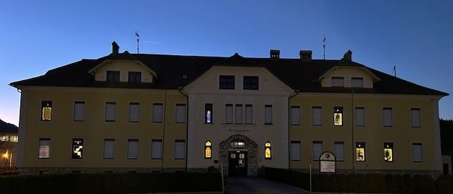 Ein großes gelbes Gebäude mit schwarzem Dach ist bei Einbruch der Dunkelheit beleuchtet. Es hat viele Fenster und einen gewölbten Eingang. Das Gebäude ist von einer Hecke und einer gut beleuchteten Einfahrt umgeben.