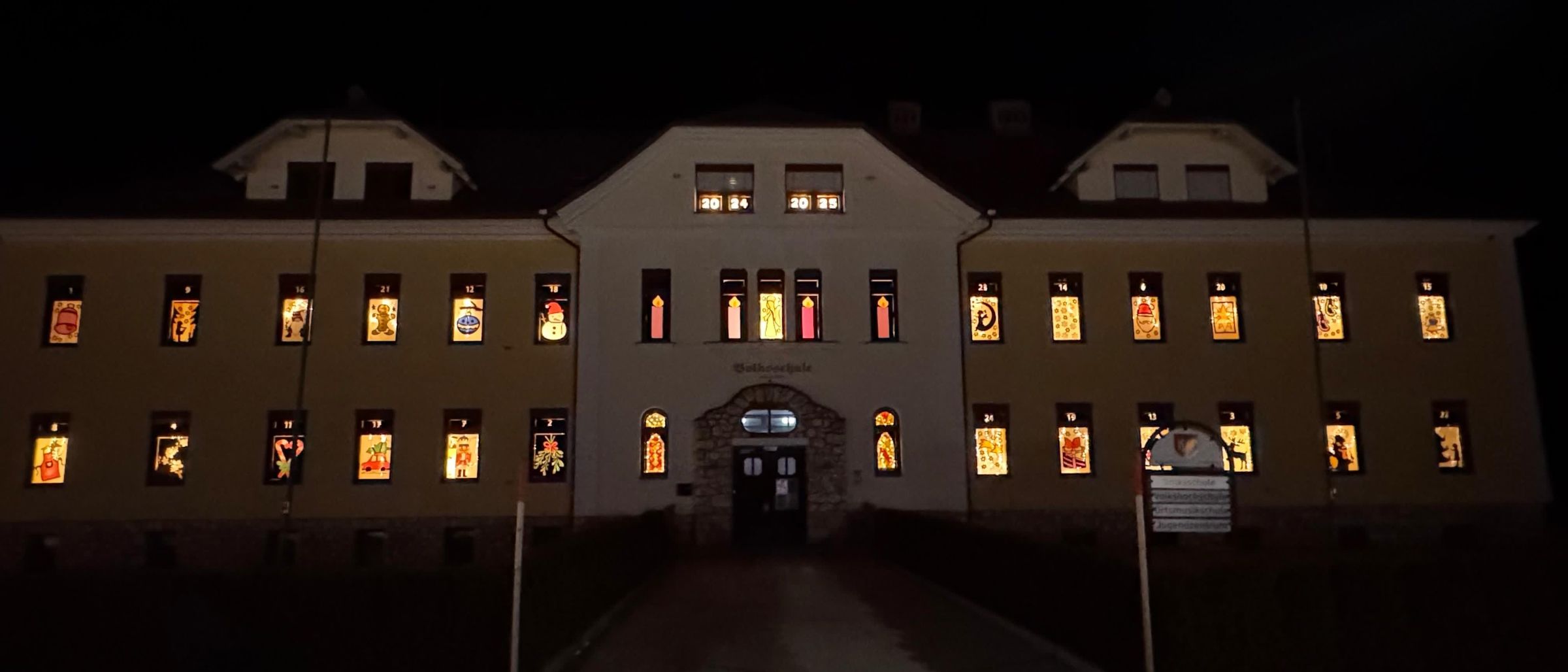 Ein Gebäude mit mehreren Fenstern, jedes mit einem anderen farbenfrohen Design, bei Nacht beleuchtet. Ein Schild steht vor dem Eingang.