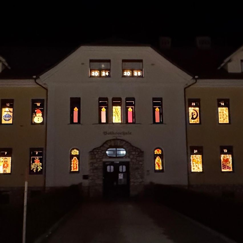 Ein Gebäude mit mehreren Fenstern, jedes mit einem anderen farbenfrohen Design, bei Nacht beleuchtet. Ein Schild steht vor dem Eingang.