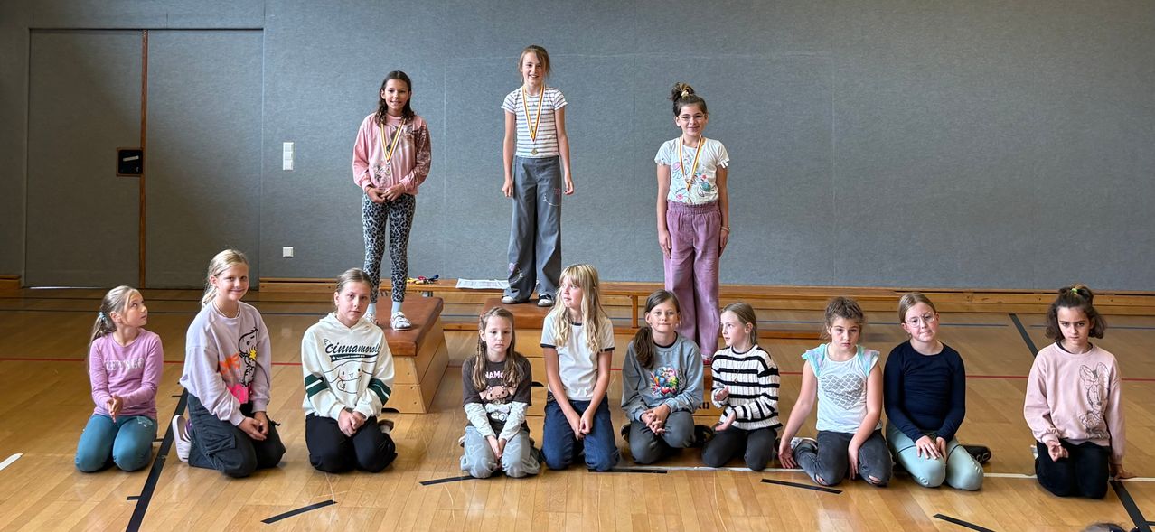 Eine Gruppe junger Mädchen, einige stehen auf hölzernen Stufen und andere sitzen auf dem Boden, befinden sich wahrscheinlich in einer Turnhalle für eine Veranstaltung, tragen Medaillen und lächeln für ein Foto.
