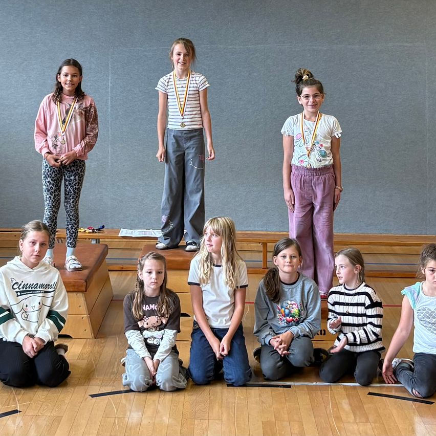 Eine Gruppe junger Mädchen, einige stehen auf hölzernen Stufen und andere sitzen auf dem Boden, befinden sich wahrscheinlich in einer Turnhalle für eine Veranstaltung, tragen Medaillen und lächeln für ein Foto.