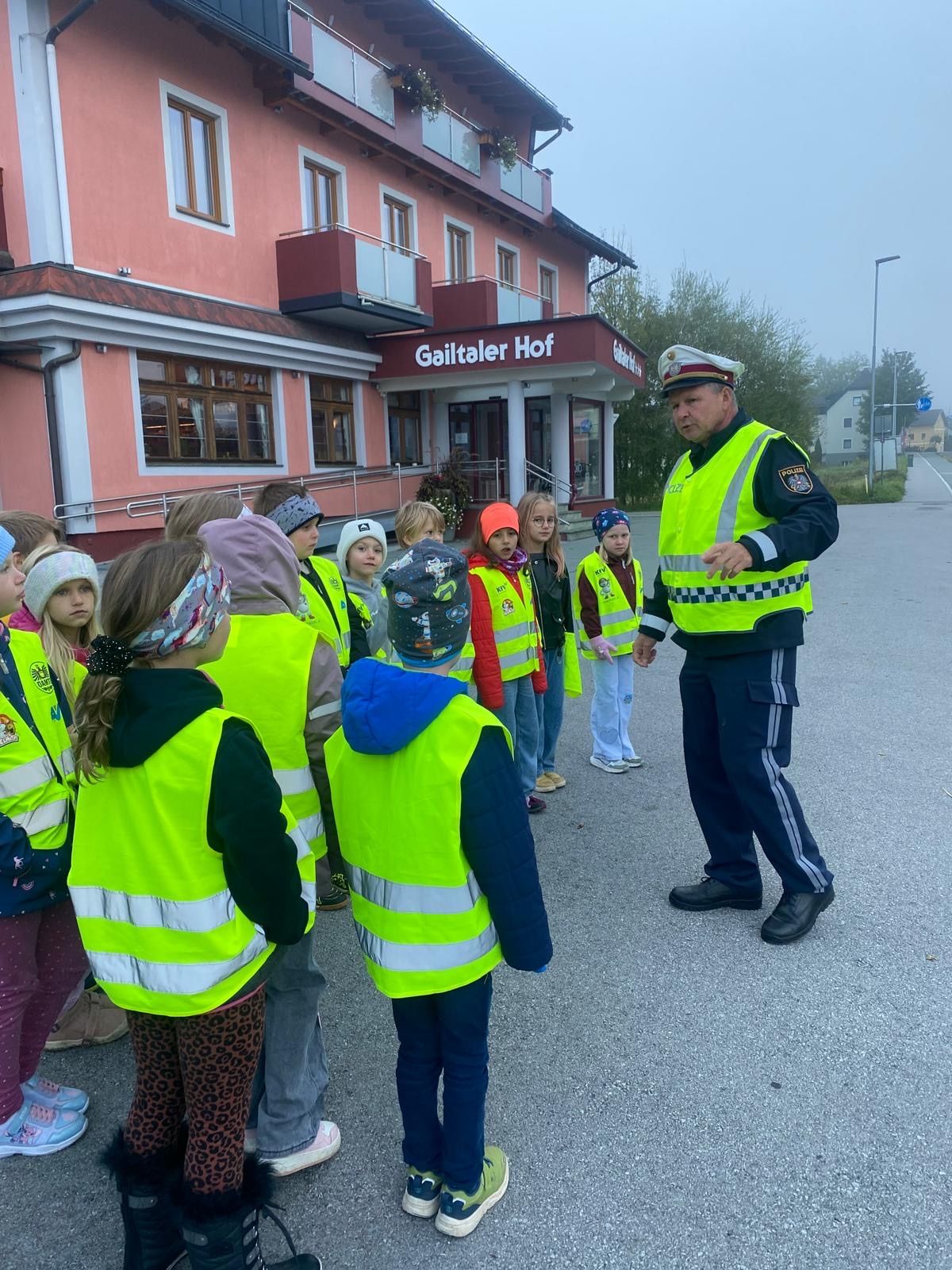 Ein Polizist in Uniform unterrichtet eine Gruppe von Kindern in Reflektorwesten vor einem rosa Gebäude mit der Aufschrift Gailtaler Hof.