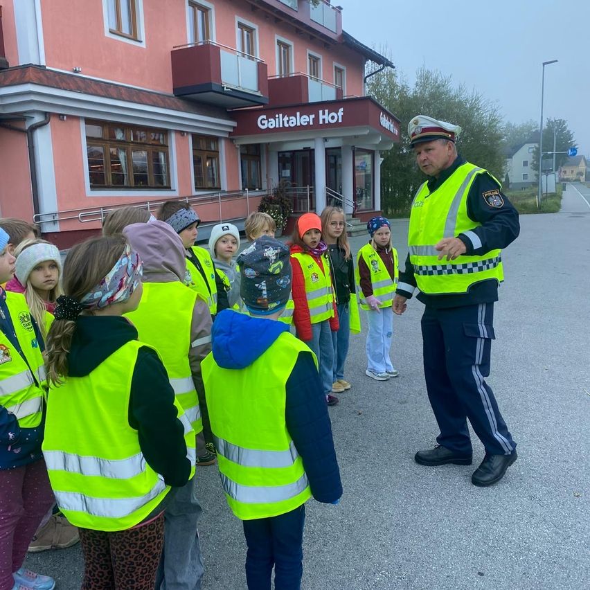 Ein Polizist in Uniform unterrichtet eine Gruppe von Kindern in Reflektorwesten vor einem rosa Gebäude mit der Aufschrift Gailtaler Hof.