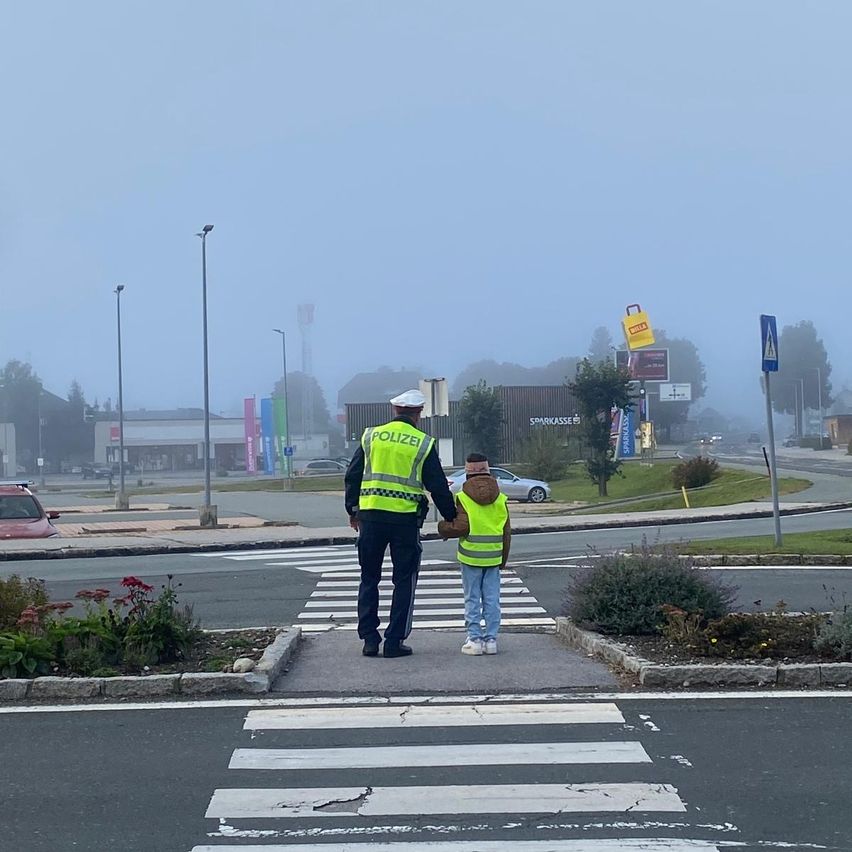 Ein Polizeibeamter und ein Kind in Warnwesten überqueren eine Straße an einem Zebrastreifen. Im Hintergrund sind Nebel und Gebäude zu sehen.