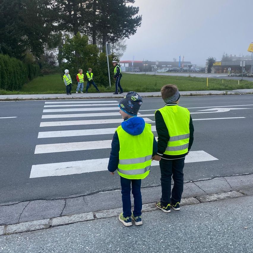 Zwei Kinder in Reflektorwesten stehen auf einem Zebrastreifen. Dahinter warten drei weitere Kinder, ebenfalls in Reflektorwesten, auf dem Bürgersteig. Ein Mann in Polizeiuniform ist in der Nähe.