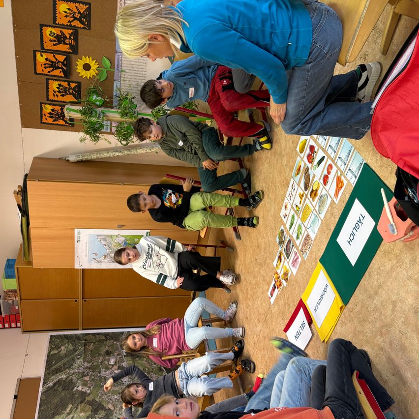 Eine Gruppe von Kindern und einem Erwachsenen sitzen im Kreis und beschäftigen sich mit einer Aktivität mit Karten auf dem Boden. Der Erwachsene ist gebeugt und hilft den Kindern. Der Raum ist mit Postern und einer Karte an der Wand dekoriert.