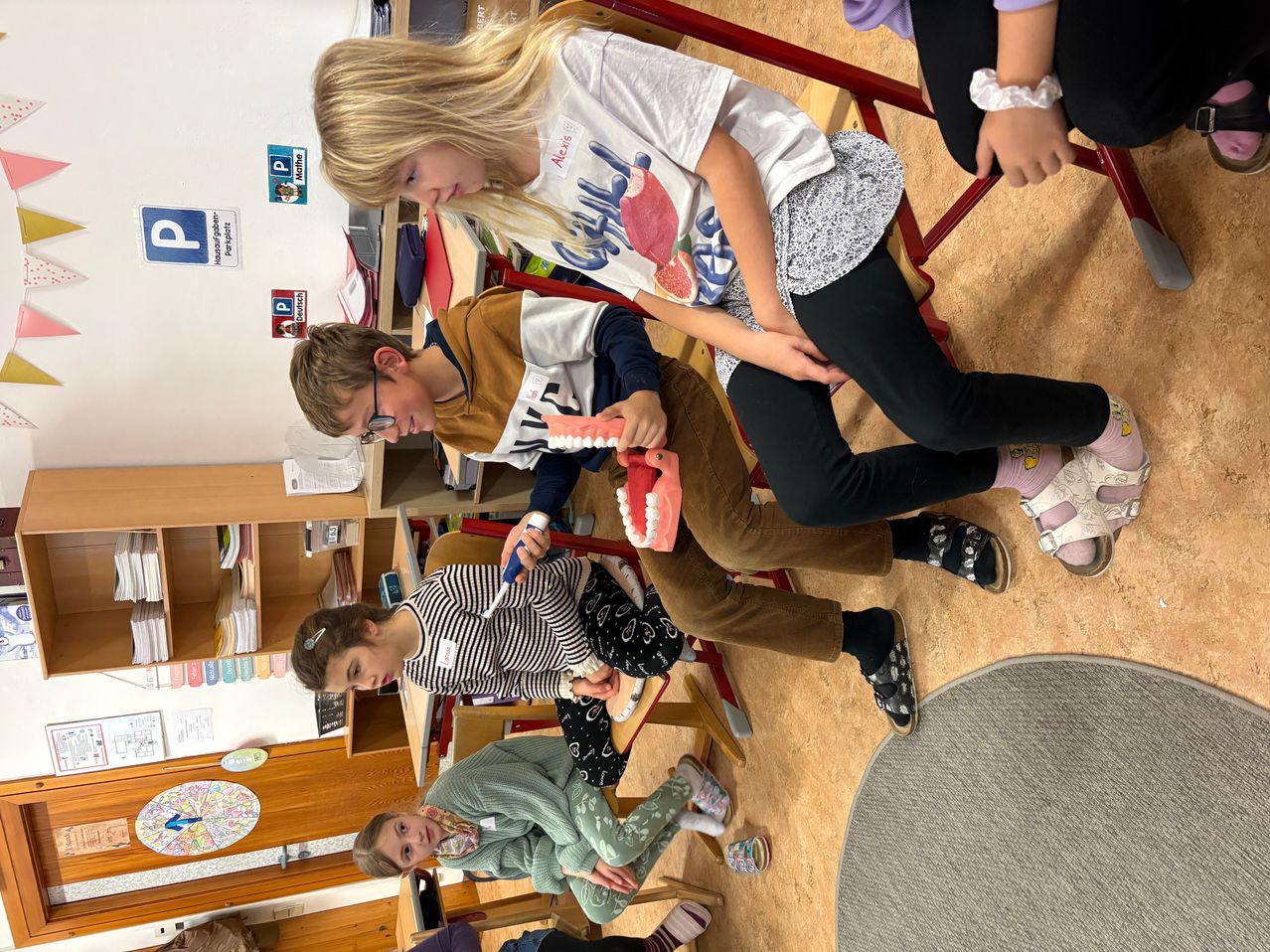 In einem Klassenzimmer versammeln sich mehrere Kinder um einen Tisch und beschäftigen sich mit einer Aktivität. Ein Mädchen hält ein Zahnmodell, während ein Junge neben ihr eine Zahnbürste benutzt. Ein weiteres Kind sitzt in der Nähe, und eine Frau im Hintergrund beobachtet.