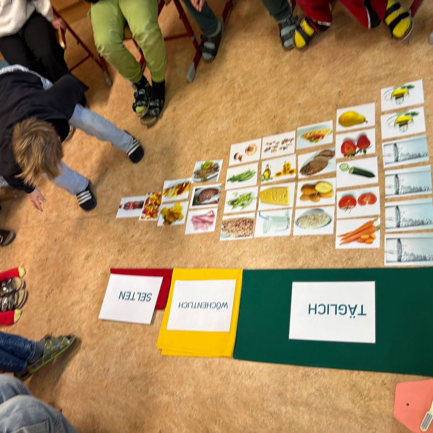 Eine Gruppe von Kindern ist auf dem Teppichboden, einige von ihnen sitzen auf Stühlen. Ein Junge in einem schwarzen Jackett ist auf den Knien. Es gibt Karten mit Bildern von Lebensmitteln auf dem Boden.