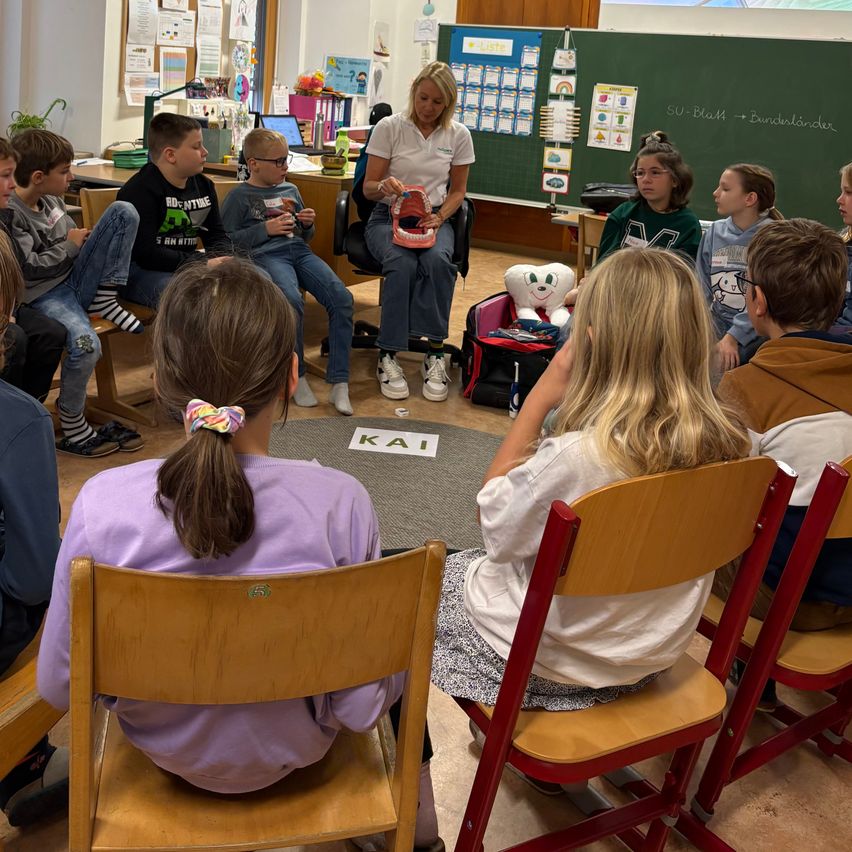 In einem Klassenzimmer sitzt eine Frau in einem Stuhl und hält ein Zahnmodell, umgeben von aufmerksamen Kindern in einem Kreis, mit einer Tafel und verschiedenen Postern im Hintergrund.