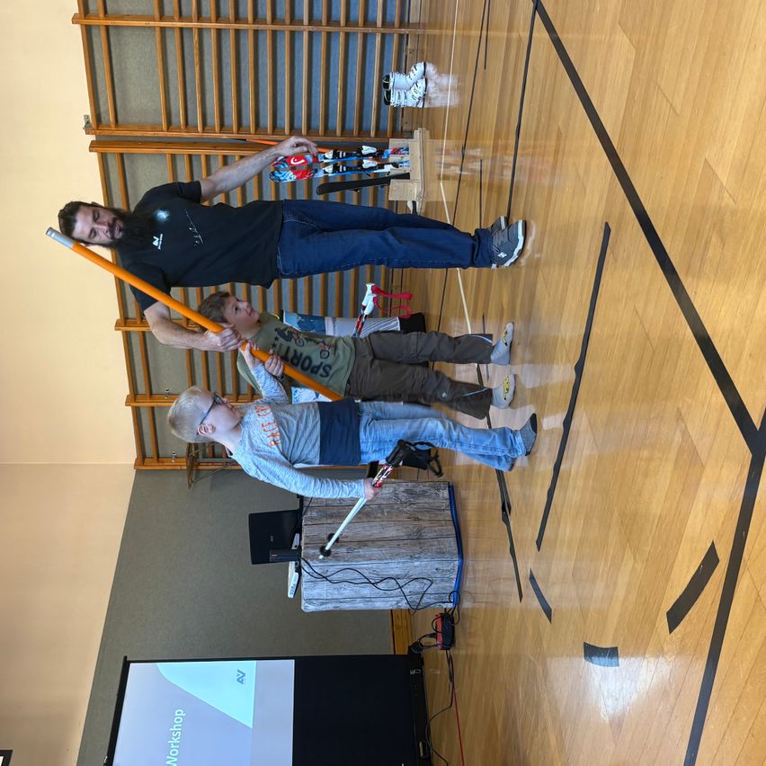 Ein Mann steht mit zwei Kindern, die lange Stöcke halten, auf einem Sporthallenboden. Sie scheinen zu üben. Der Boden hat Markierungen. Ein hölzerner Podest steht vor einer Projektionsleinwand.