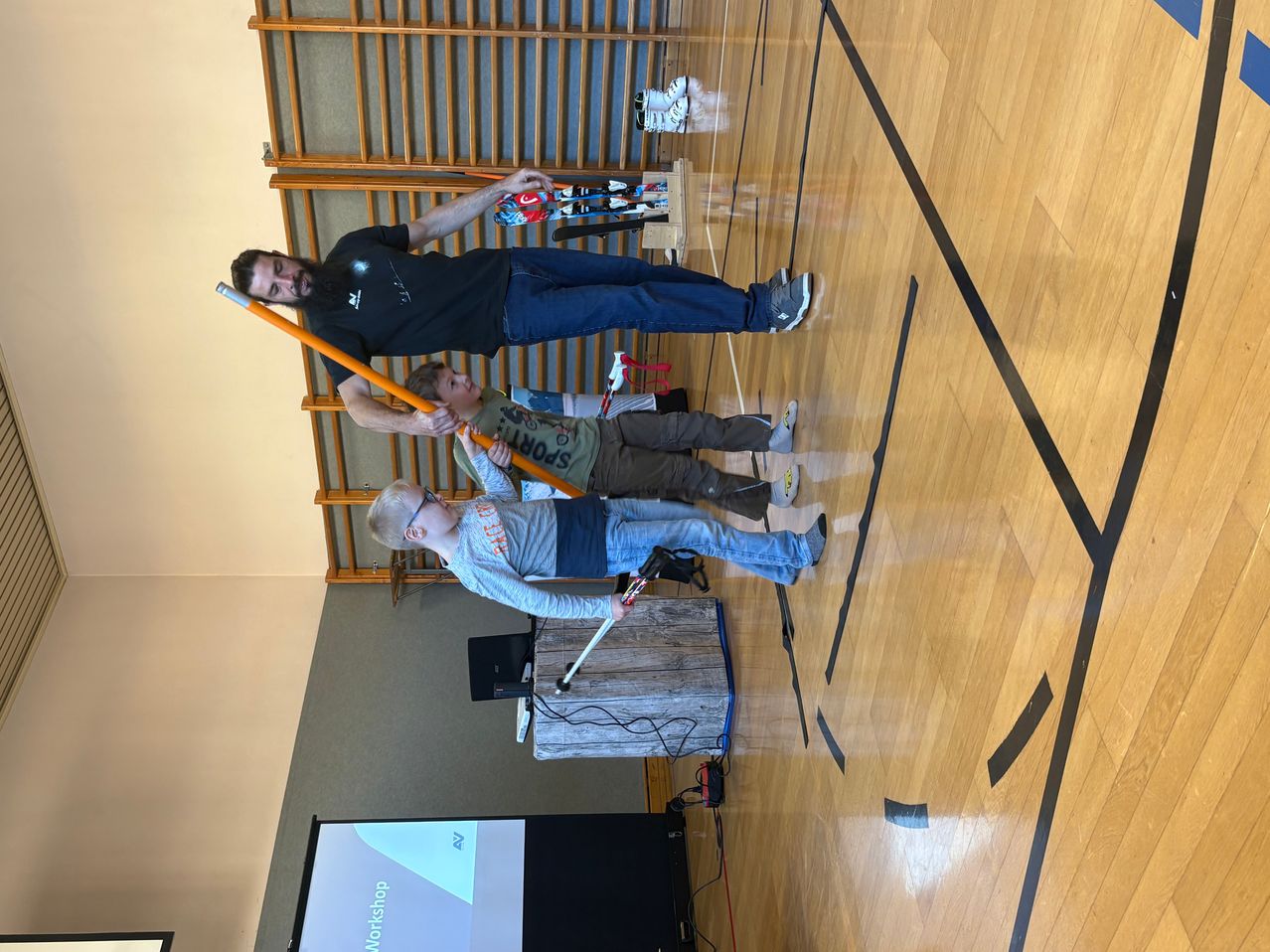 Ein Mann steht mit zwei Kindern, die lange Stöcke halten, auf einem Sporthallenboden. Sie scheinen zu üben. Der Boden hat Markierungen. Ein hölzerner Podest steht vor einer Projektionsleinwand.