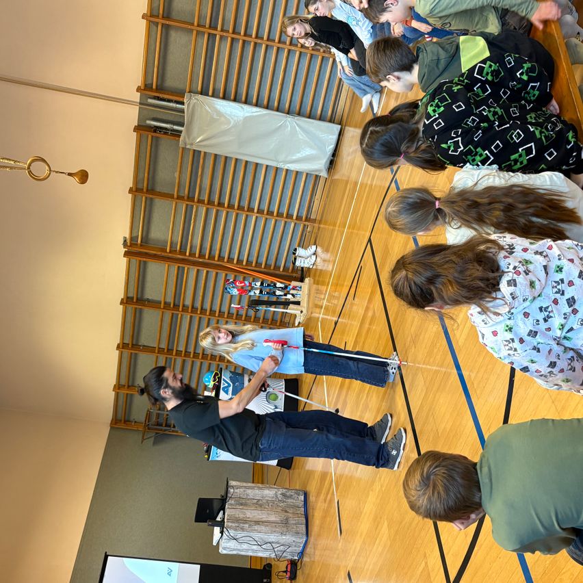 Eine Gruppe von Kindern und Erwachsenen versammelt sich in einer Turnhalle. Sie schauen alle auf einen Mann mit Mikrofon und Stock, der etwas demonstriert. Die Kinder halten Stöcke und stehen in einer Reihe.