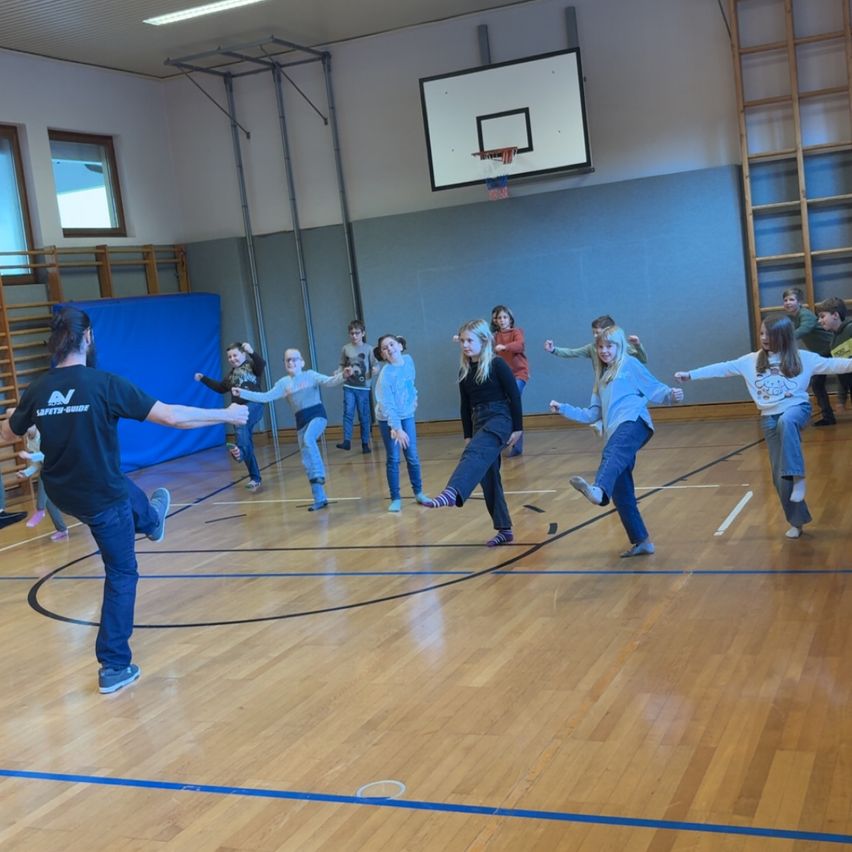 In einer Turnhalle spielen eine Gruppe von Kindern ein Spiel, während ein Mann sie anführt. Sie springen und haben Spaß.