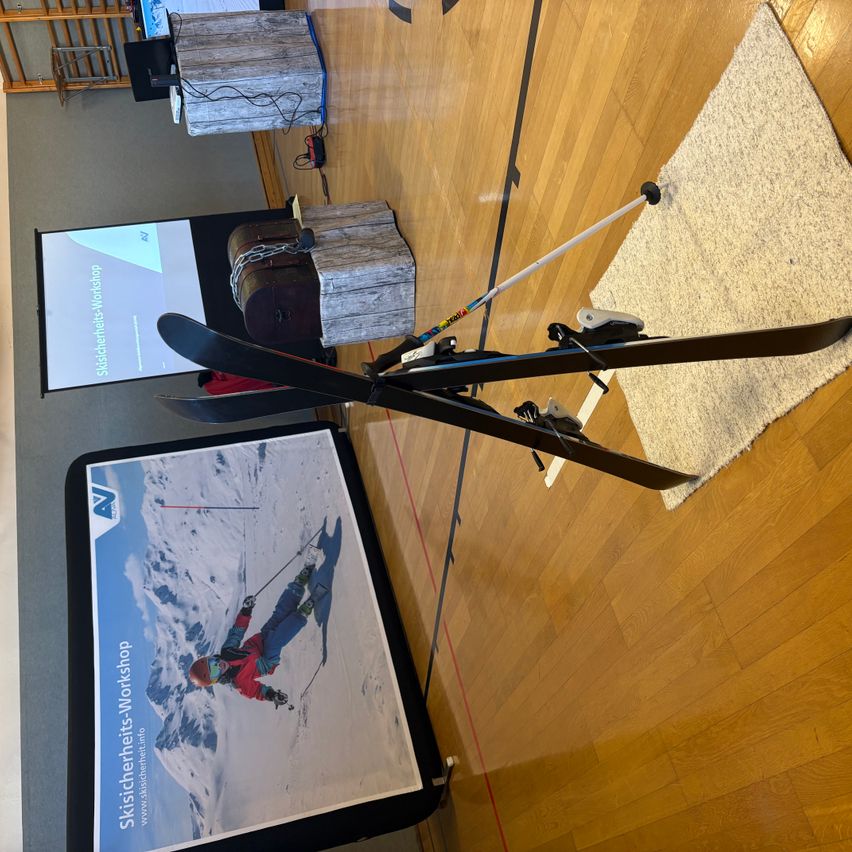 Ein Bild zeigt einen Ski mit angebundenen Skischuhen, der auf einem Holzboden neben einem Poster liegt, das einen Skisicherheits-Workshop bewirbt. Der Boden ist mit einem weißen Teppich bedeckt. Im Hintergrund befindet sich ein Präsentationsbildschirm mit dem Text 'Skisicherheits-Workshop'.
