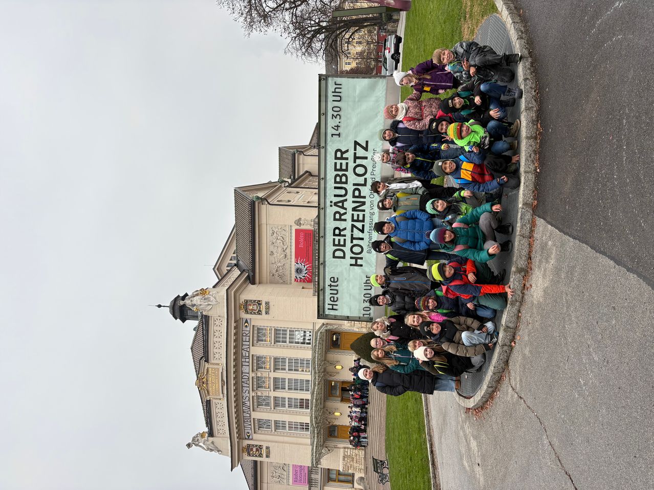 Eine Gruppe von Menschen posiert vor einem großen Banner vor einem historischen Gebäude. Das Banner trägt in fetten Buchstaben 'Der Räuber Hotzenplotz'.
