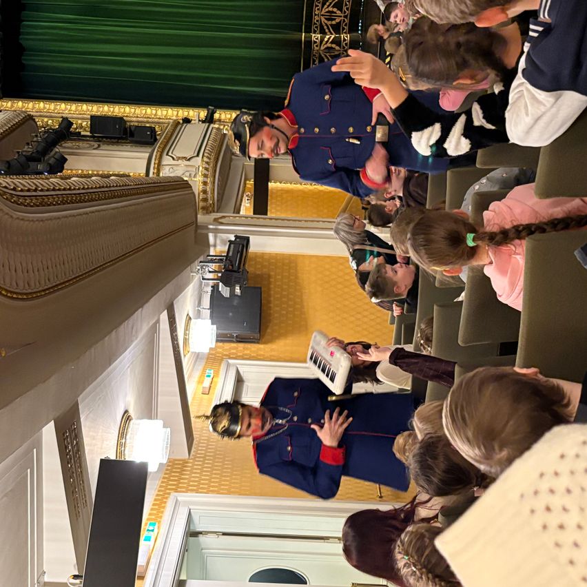 Eine Gruppe von Kindern sieht sich in einem Theater eine Aufführung an, angeführt von einem Mann in einer blauen Uniform, der eine Tastatur spielt.