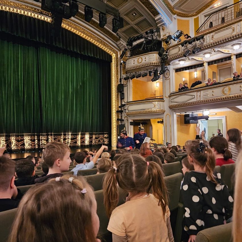 Ein Theater mit grünem Vorhang und gelben Wänden. Kinder sitzen im Publikum und beobachten eine Aufführung, mit einigen Erwachsenen auf der Galerie.