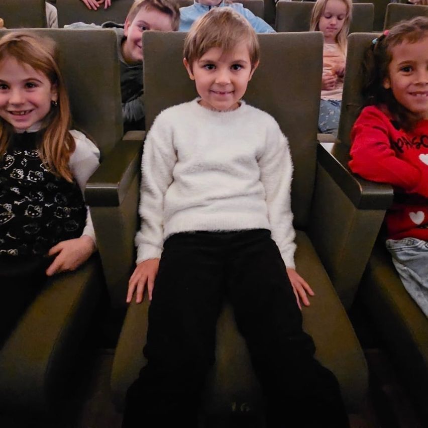 Mehrere Kinder sitzen in einem Theater und lächeln. Ein Junge in einem weißen Pullover und schwarzen Hosen sitzt in der Mitte, während andere rote und schwarze Kleidung tragen.