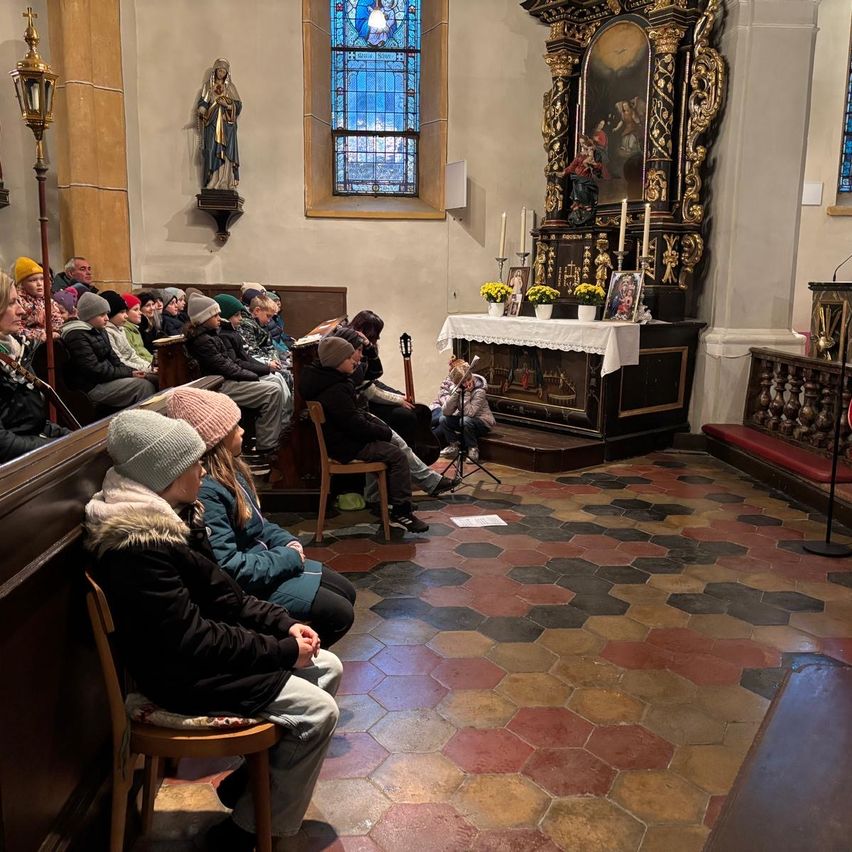 Eine Gruppe von Menschen sitzt in einer Kirche. Eine Person steht mit einer Gitarre vor ihnen. Kerzen stehen auf dem Altar, und eine Statue ist an der Wand befestigt.