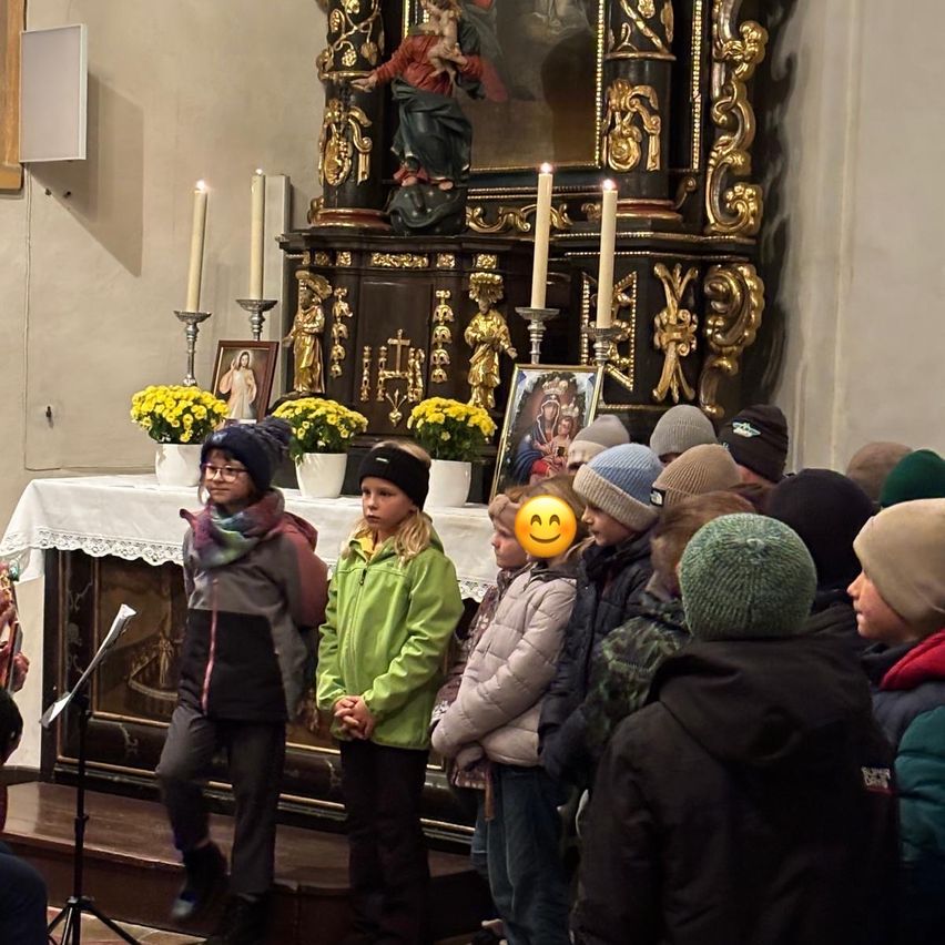 In einer Kirche stehen mehrere Kinder in einer Reihe und tragen Winterkleidung. Vor ihnen steht ein Altar mit einer Statue, Blumen und brennenden Kerzen. Eine Person steht hinter einem Mikrofon.
