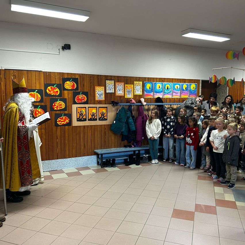 Eine als König verkleidete Person liest aus einem Buch in einem Klassenzimmer, mit Kindern, die sich versammelt haben, einige stehen vor einem Kleiderständer, an dem verschiedene Jacken hängen.