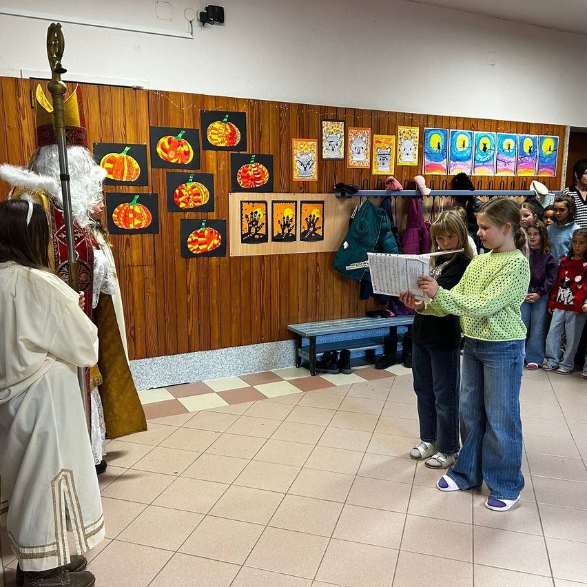 Eine Gruppe von Kindern steht in einem Flur, eines hält eine Musikpartitur in der Hand, während ein anderes in einem Kostüm steht. Die Wand ist mit verschiedenen Kunstwerken dekoriert.