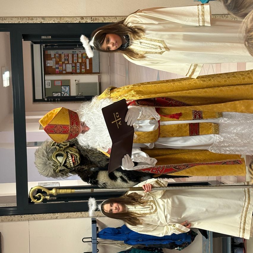 Eine Gruppe von Menschen in Kostümen. Ein Mann, der als Bischof gekleidet ist, hält ein Buch. Eine Frau in einem weißen Gewand steht hinter ihm. Eine andere Frau in einem weißen Gewand steht davor.