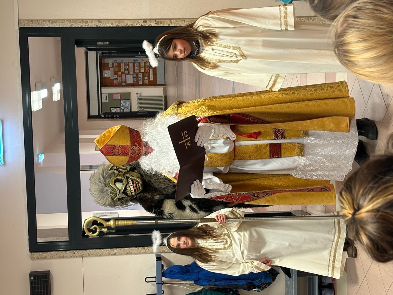 Eine Gruppe von Menschen in Kostümen. Ein Mann, der als Bischof gekleidet ist, hält ein Buch. Eine Frau in einem weißen Gewand steht hinter ihm. Eine andere Frau in einem weißen Gewand steht davor.