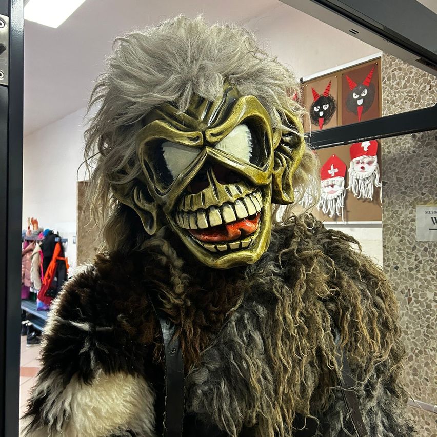 Eine Person trägt eine goldene Maske mit einem gruseligen Gesicht in einem Raum. Die Maske hat ein Schädeldesign. Die Person trägt ein pelziges Kostüm.