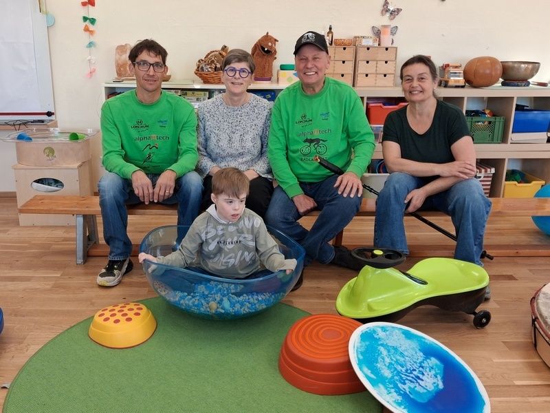 Fünf Menschen, einschließlich eines Jungen, sitzen auf einer Bank in einem Spielzimmer. Vier Erwachsene tragen grüne Shirts und der Junge ein graues Sweatshirt. Sie alle sehen glücklich aus.