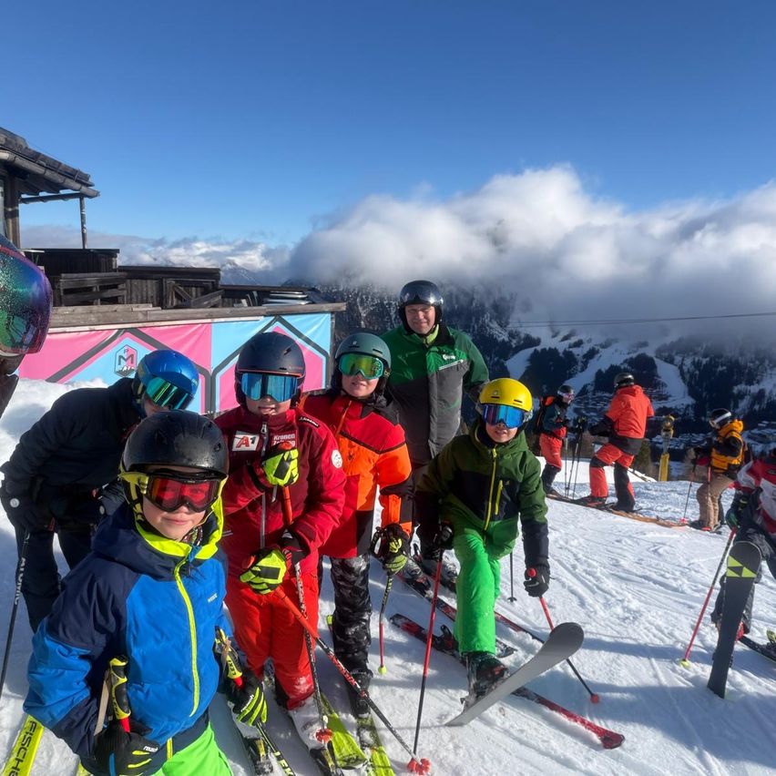 Mehrere Personen stehen auf einem verschneiten Hang mit Skiern und tragen Winterkleidung und Helme. Ein Kind in einer grünen Jacke hält Skistöcke. Dahinter steht ein Gebäude mit einer rosa-blauen Wand.