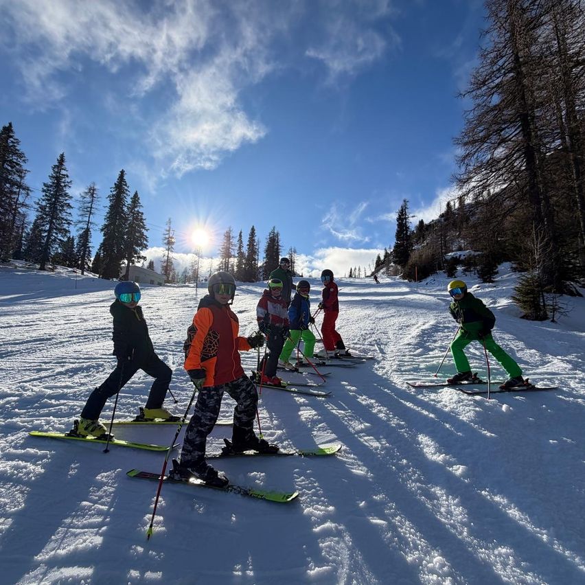 Eine Gruppe junger Skifahrer, ausgestattet mit Helmen und Skibrillen, fährt den schneebedeckten Hang hinunter. Der Himmel ist blau mit einigen Wolken, und Bäume sind im Hintergrund zu sehen.