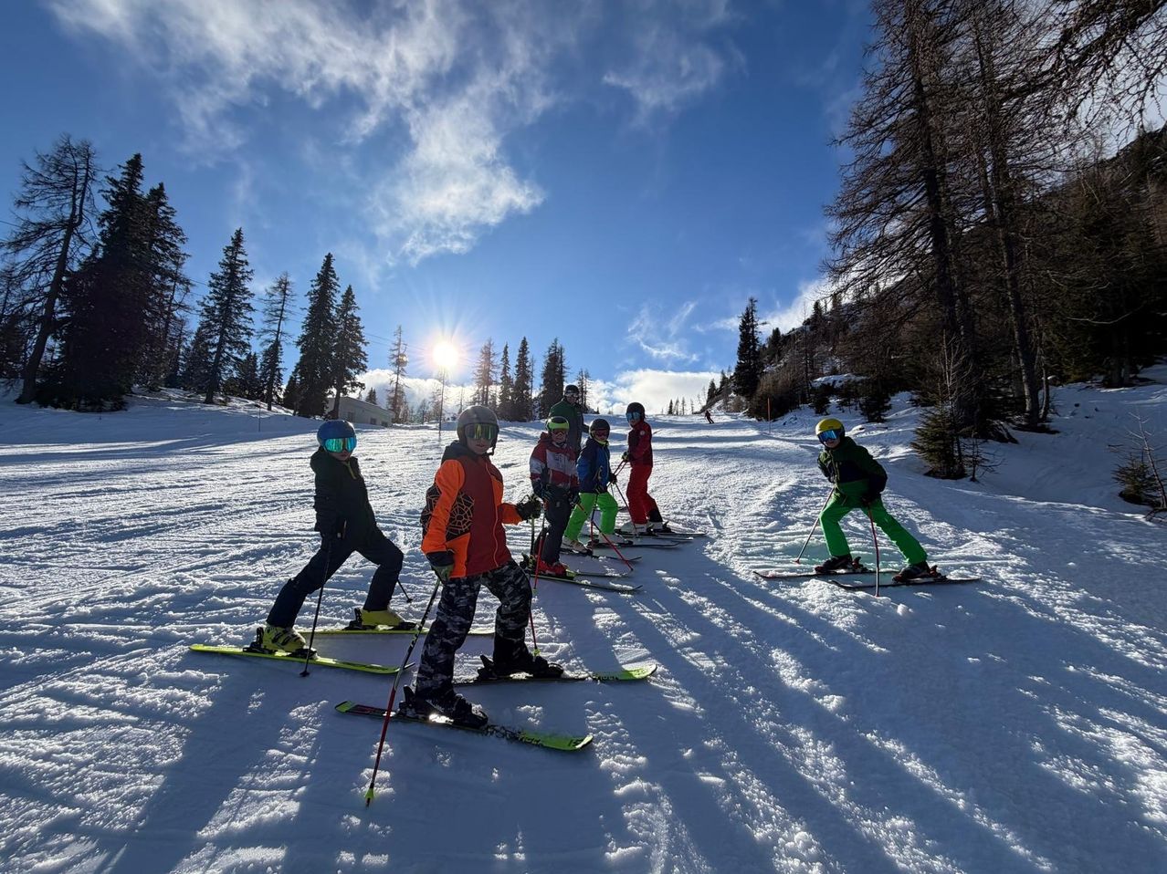 Eine Gruppe junger Skifahrer, ausgestattet mit Helmen und Skibrillen, fährt den schneebedeckten Hang hinunter. Der Himmel ist blau mit einigen Wolken, und Bäume sind im Hintergrund zu sehen.
