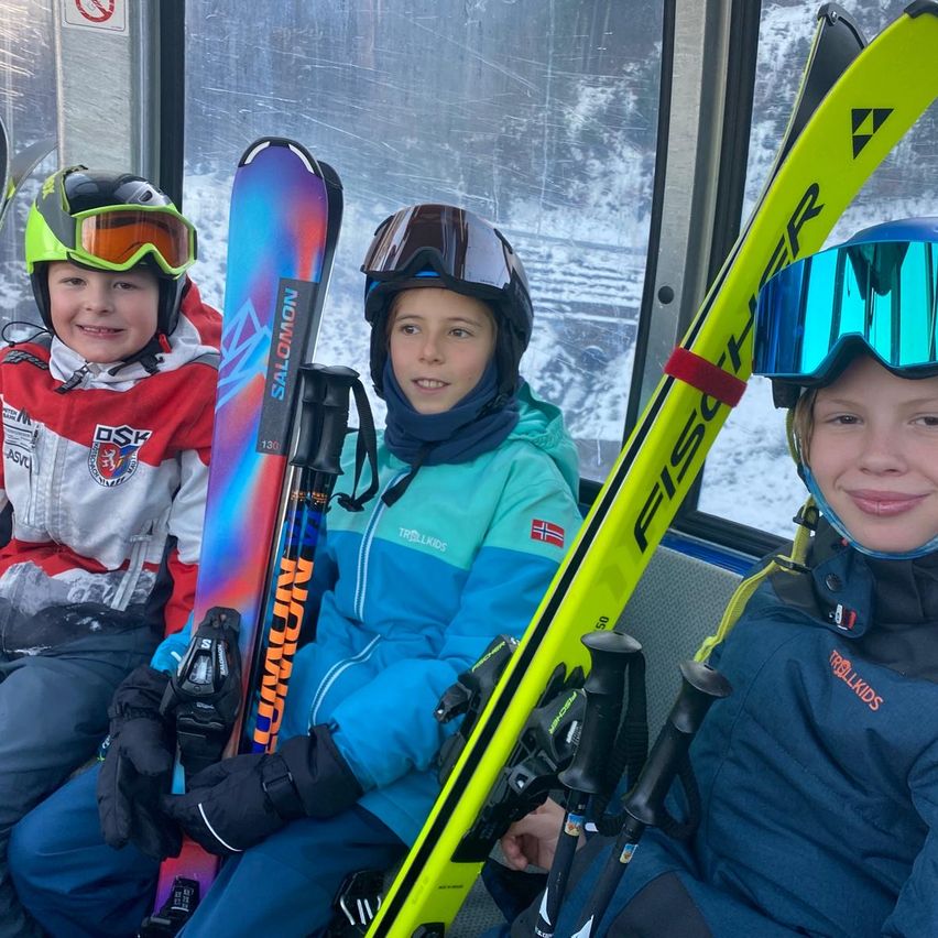 Drei Kinder, alle in Winterkleidung, sitzen mit Skiern in einer Gondel. Zwei von ihnen halten Skistöcke, während das dritte Kind die Skier auf dem Schoß hat. Sie alle lächeln in die Kamera.