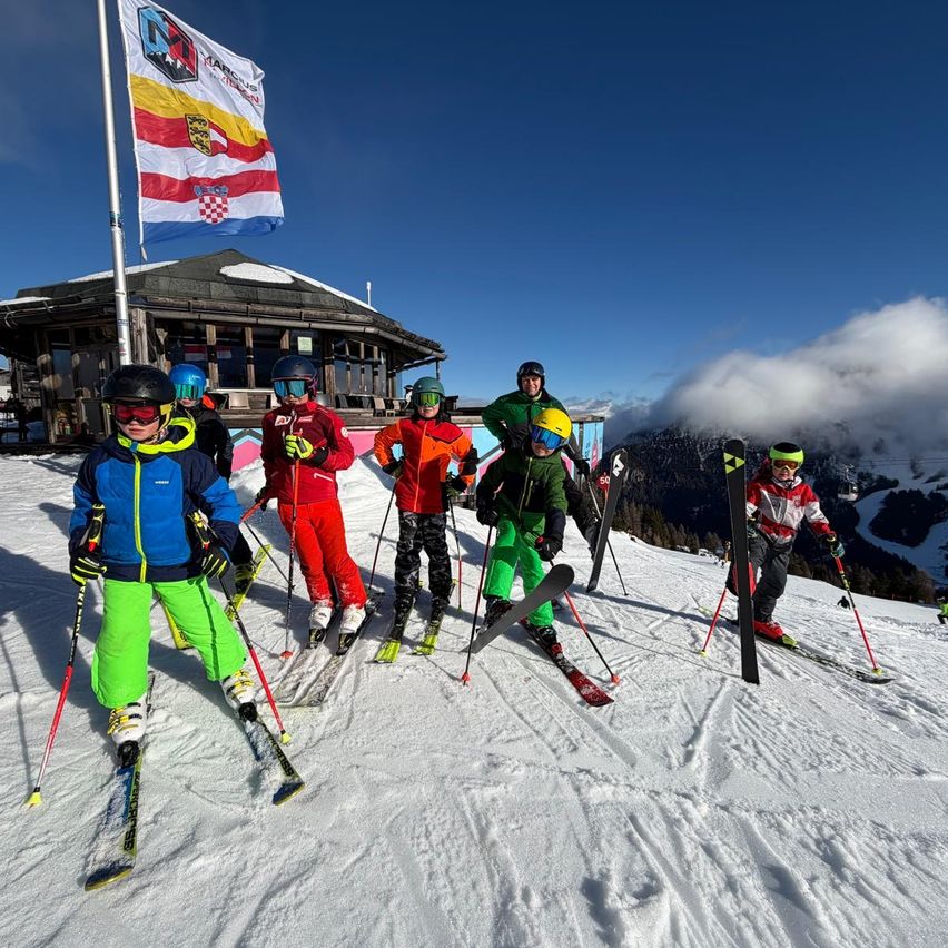 Eine Gruppe von Skifahrern in bunten Skianzügen posiert für ein Foto auf einer verschneiten Piste, mit einer Flagge und einem Gebäude im Hintergrund.