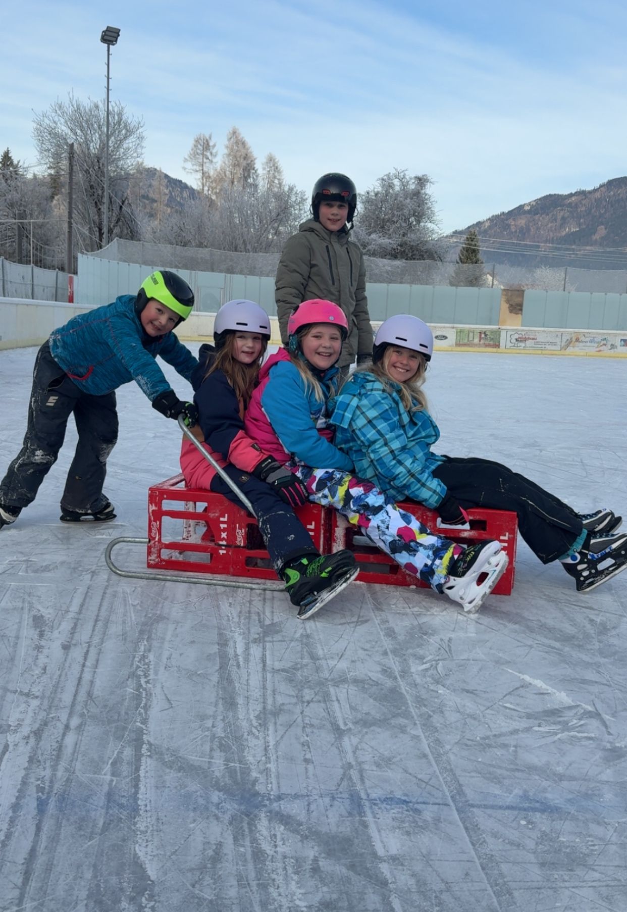 Fünf Kinder auf einer Eisbahn, in Winterkleidung und mit Helmen. Drei sitzen auf einem roten Schlitten, während zwei ziehen. Berge und Bäume sind im Hintergrund.