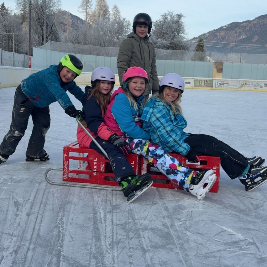 Fünf Kinder auf einer Eisbahn, in Winterkleidung und mit Helmen. Drei sitzen auf einem roten Schlitten, während zwei ziehen. Berge und Bäume sind im Hintergrund.