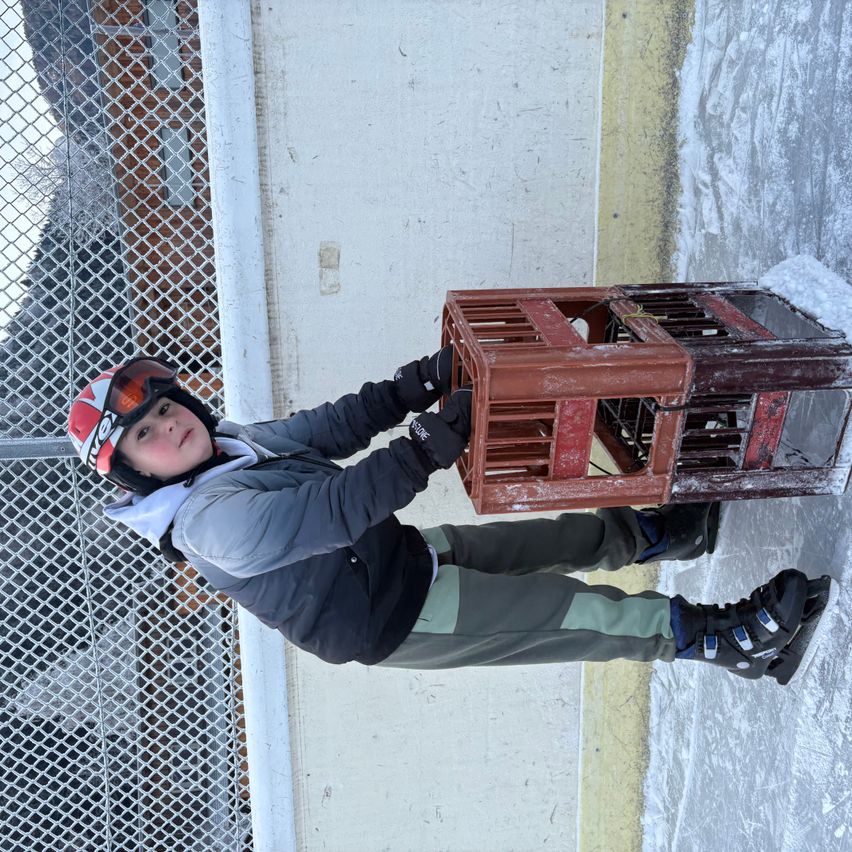 Ein junger Mensch in Winterkleidung hält einen roten Kasten und steht auf Eis, in der Nähe eines Zauns.