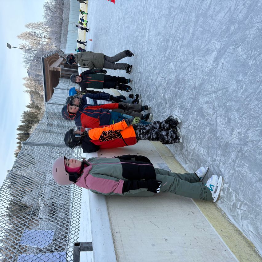 Eine Gruppe von Menschen steht auf dem Eislaufplatz, einige von ihnen tragen Helme und Schlittschuhe. Die Eisbahn ist von einem Zaun umgeben. In der Ferne sind Bäume und ein Gebäude zu sehen.