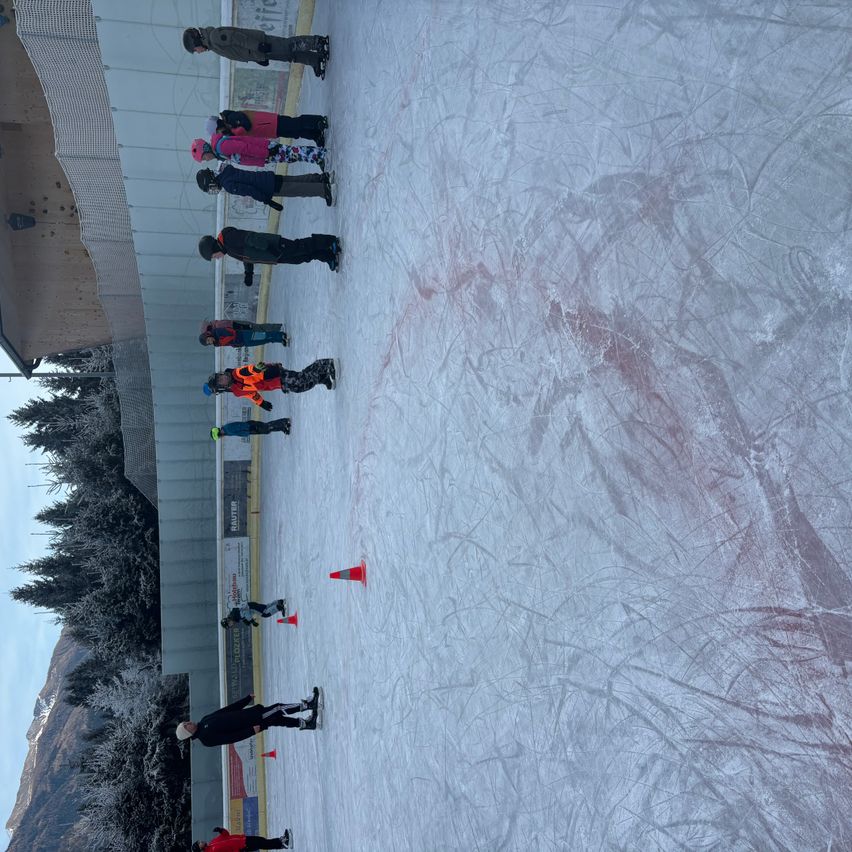 Eine Eisbahn im Freien, auf der mehrere Menschen in Winterkleidung stehen. Einige halten Stöcke. Orangefarbene Kegel markieren die Ränder der Eisbahn, und schneebedeckte Bäume und ein Gebäude sind im Hintergrund zu sehen.