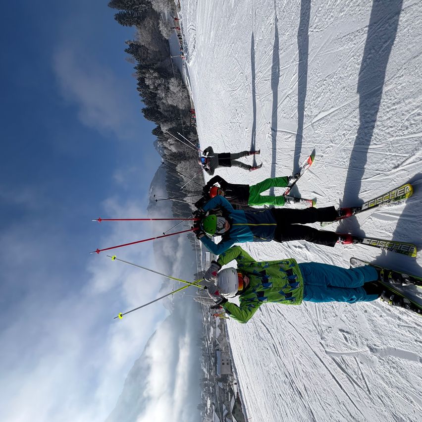 Eine Gruppe von Skifahrern in bunter Kleidung fährt einen verschneiten Hang hinunter unter einem blauen Himmel mit verstreuten Wolken.