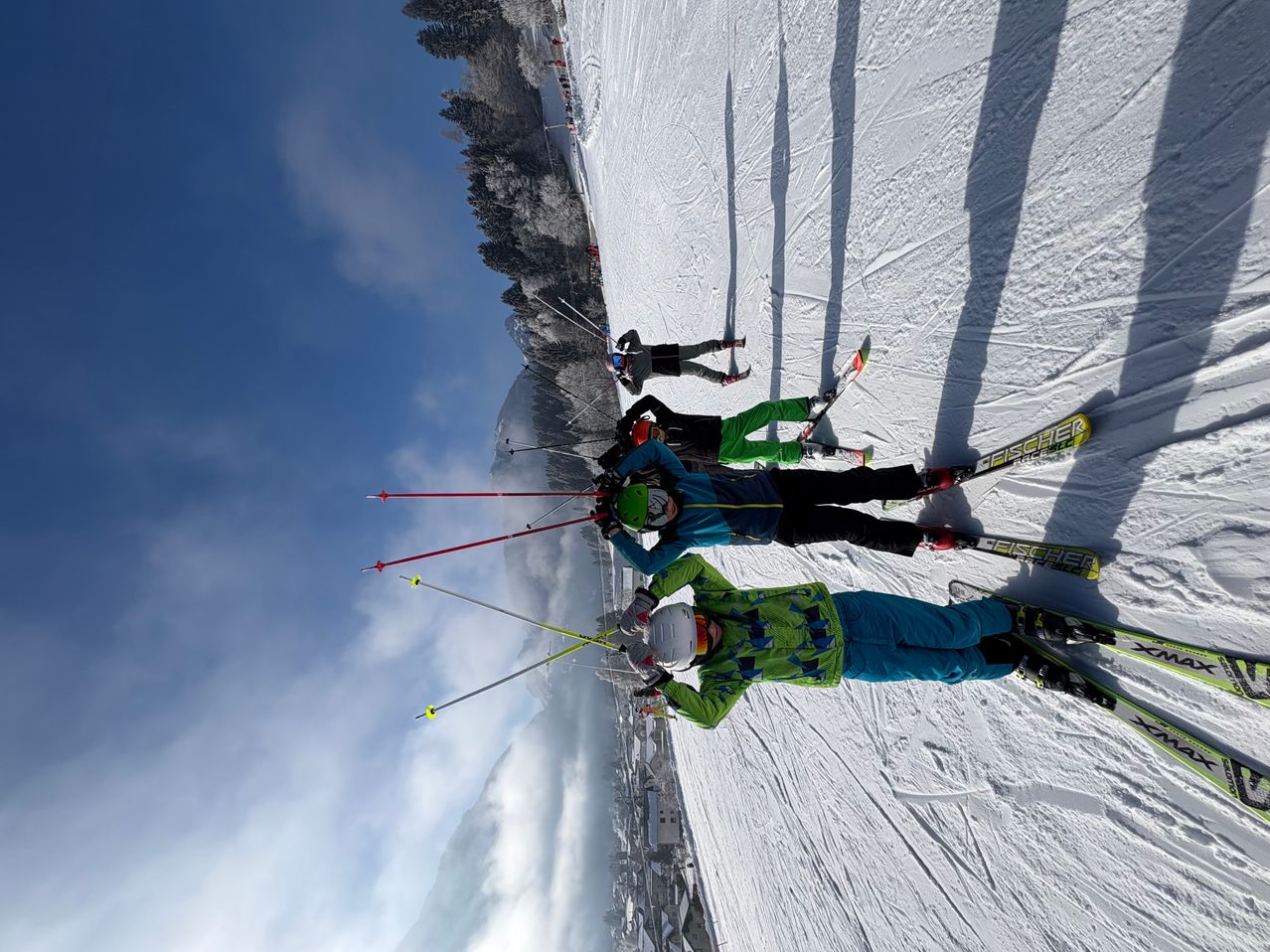 Eine Gruppe von Skifahrern in bunter Kleidung fährt einen verschneiten Hang hinunter unter einem blauen Himmel mit verstreuten Wolken.