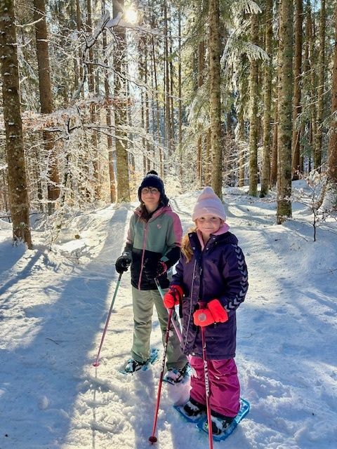 Zwei Kinder in Winterkleidung fahren durch einen verschneiten Wald. Sie tragen Skier und halten Skistöcke. Der Boden und die Bäume sind schneebedeckt.