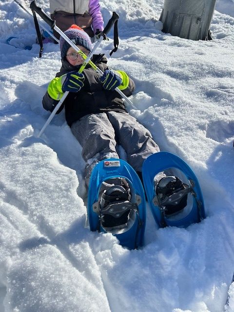 Ein Kind in Schneeschuhen liegt auf dem Schnee, während ein anderes Kind Skistöcke im Hintergrund hält.