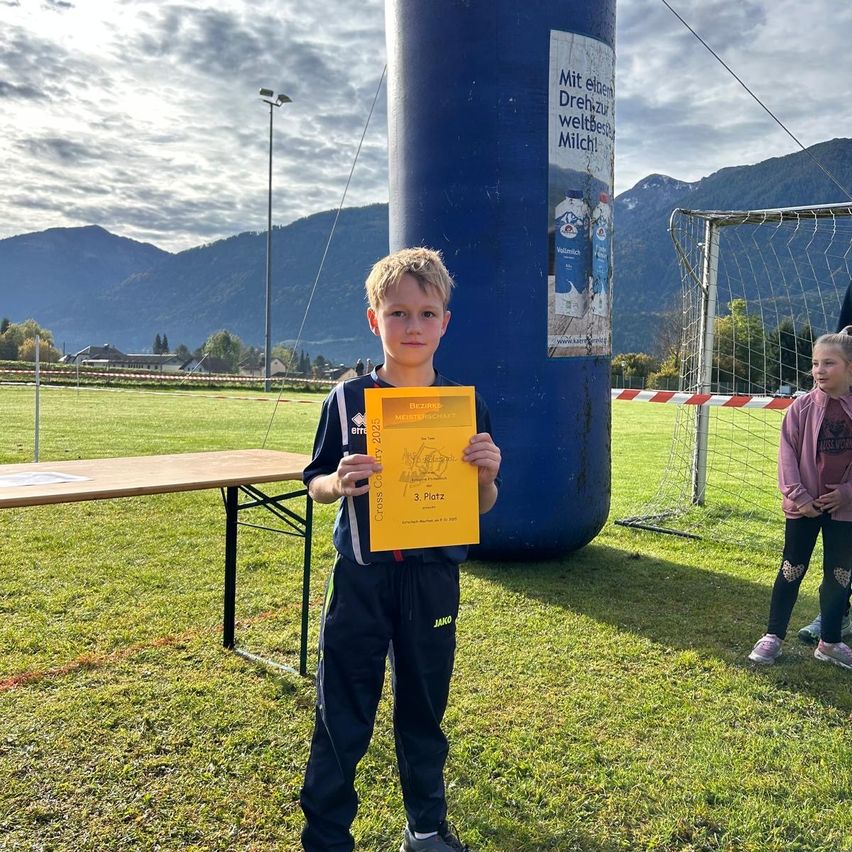 Ein junger Junge steht auf einem Rasenfeld und hält ein gelbes Zertifikat in der Hand, möglicherweise bei einer Sportveranstaltung. Hinter ihm steht ein Fußballtor. In der Ferne sind Berge sichtbar. Ein Tisch mit einem weißen Papier darauf ist ebenfalls in der Szene.