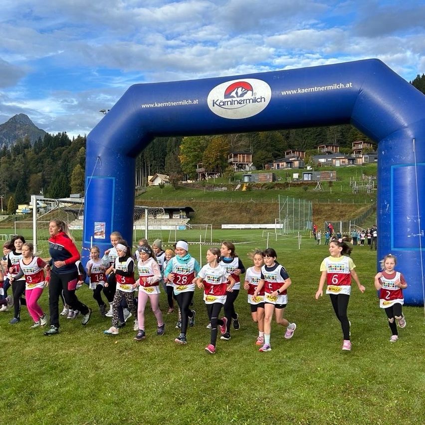 Eine Gruppe junger Mädchen in nummerierten Trikots läuft unter einem Bogen mit dem Namen Karntnermilch. Der Hintergrund hat ein grünrasiges Feld mit Häusern und Bäumen.