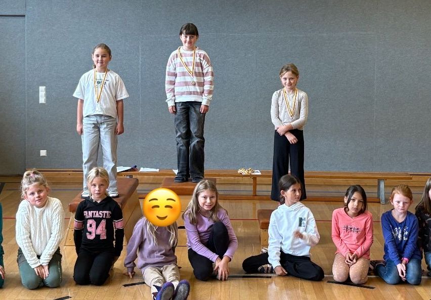 Eine Gruppe von Kindern sitzt und steht in einem Raum. Ein Kind steht auf einer Holzbank. Sie tragen alle Medaillen. Einem der Kinder wurde ein Emoji hinzugefügt.